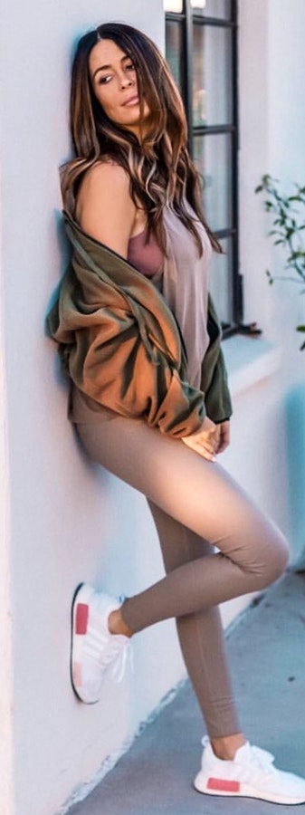 Woman wearing brown jacket and white sneakers leaning on wall.