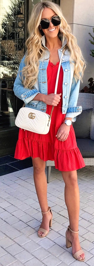 Woman in blue denim jacket and red dress holding white leather crossbody bag wearing black sunglasses.