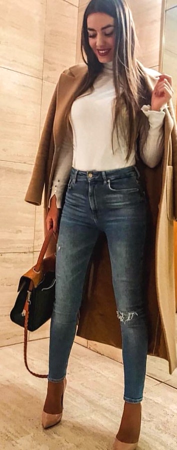 Woman in white long-sleeved shirt, distressed blue-washed jeans with beige coat outfit while holding 2-way bag.