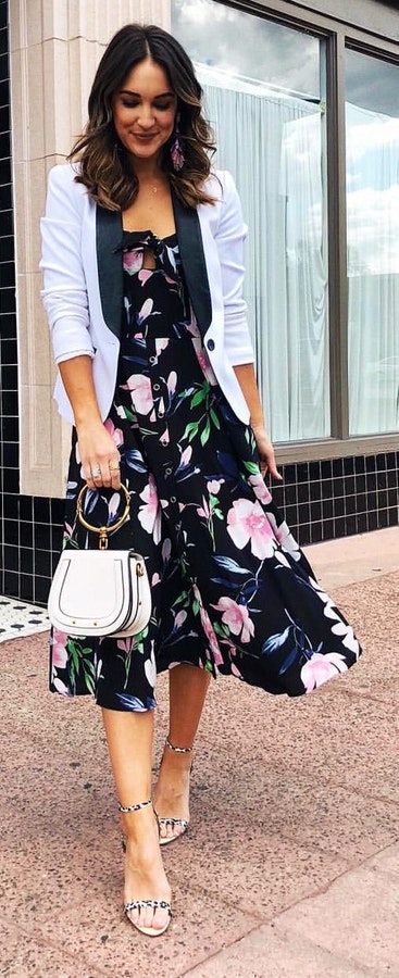 Woman wearing black floral dress and white blazer with handbag.