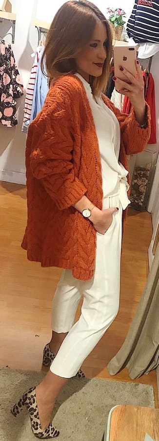 Woman wearing orange cable-knit cardigan and white fitted jeans taking mirror selfie.